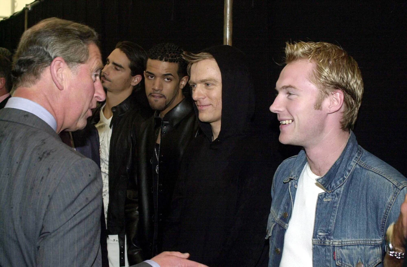 Ronan Keating (pierwszy z prawej) podczas spotkania z ówczesnym księciem Karolem, obok Keatinga Bryan Adams - Londyn, 9 lipca 2000 r. Grupa młodych mężczyzn uśmiecha się i rozmawia z mężczyzną w garniturze w jasnym świetle, tłum w tle pozostaje niewyraźny, atmosfera jest przyjazna i oficjalna.