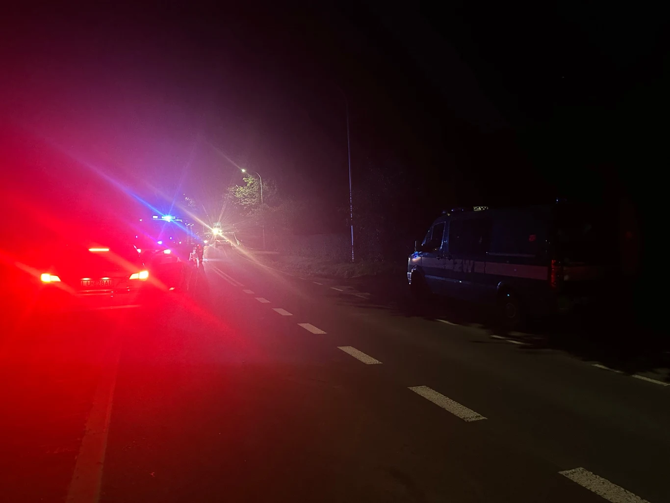 nocna scena na jezdni z czerwonymi i niebieskimi światłami radiowozów policyjnych, radiowóz oraz drugi pojazd zatrzymane na poboczu, słabo oświetlona droga, brak widocznych osób