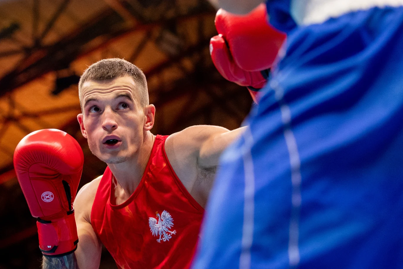 Paweł Brach Bokser w czerwonym stroju z orłem na piersi wykonuje ruch podczas walki z przeciwnikiem ubranym w niebieski strój, w tle widoczne rozmyte elementy hali sportowej.