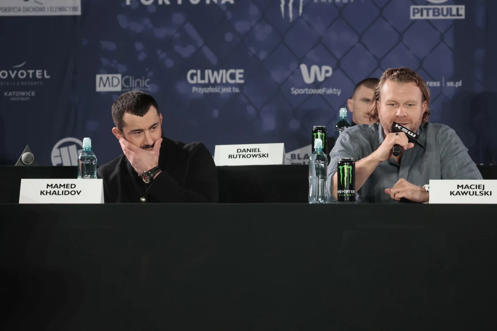 Dwóch mężczyzn siedzących przy stole konferencyjnym, przed nimi stoją butelki z wodą, mikrofon i tabliczki z nazwiskami, jeden z mężczyzn mówi do mikrofonu, drugi ma zamyśloną minę.