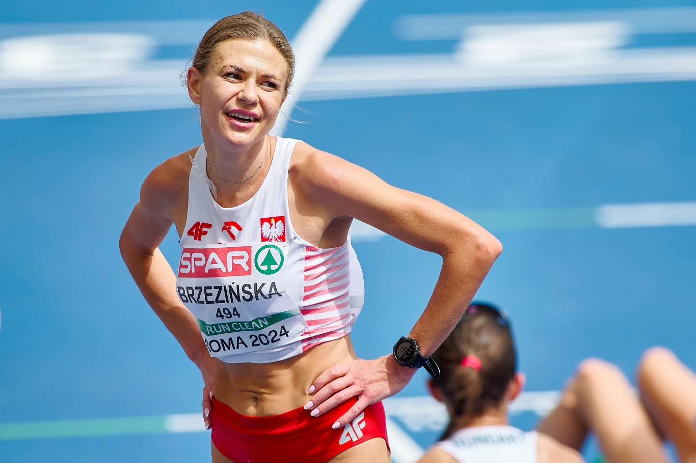 Zawodniczka reprezentująca Polskę w stroju sportowym z numerem startowym, uśmiechnięta po zakończeniu biegu na stadionie lekkoatletycznym, w tle inna sportsmenka siedzi na bieżni.