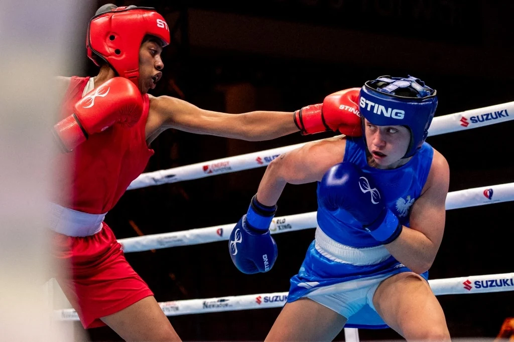 Julia Szeremeta pewna medalu MŚ Pojedynek dwóch bokserów na ringu, jeden w czerwonym stroju zadawał cios, drugi w niebieskim unikał uderzenia, obaj zawodnicy wyposażeni w kaski ochronne oraz rękawice bokserskie.