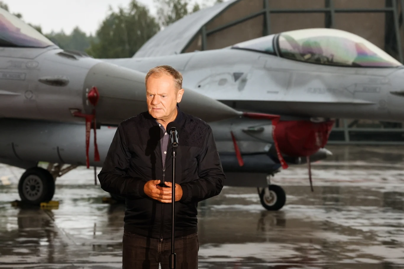 Donald Tusk odwiedził 32. Bazę Lotnictwa Taktycznego w Łasku Mężczyzna stojący przy mikrofonie na tle myśliwca wojskowego w deszczowej scenerii, obok hangaru lotniczego.