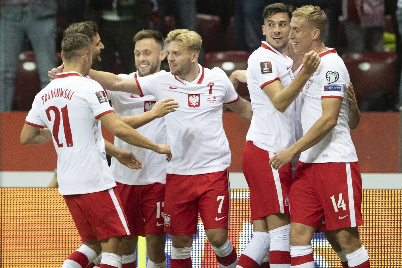 Kamil Jóźwiak (w środku) Grupa piłkarzy w biało-czerwonych strojach świętuje zdobycie gola podczas meczu piłkarskiego, wszyscy zawodnicy są uśmiechnięci i obejmują się nawzajem.