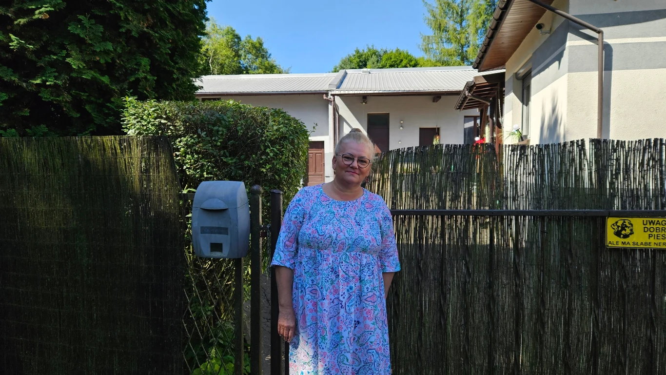 Starsza kobieta w sukience w kwiaty stoi przy ogrodzeniu przed domem jednorodzinnym, w tle krzewy i drzewa, na bramie widoczna skrzynka na listy oraz żółta tabliczka ostrzegawcza o psie.