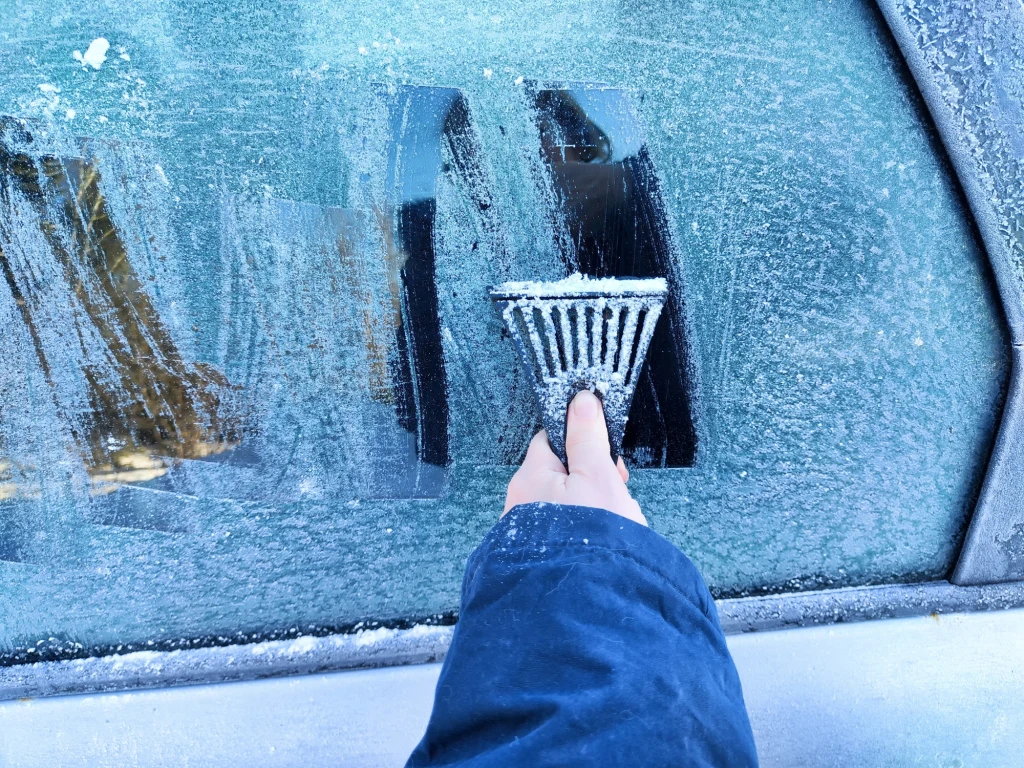 Ręka w niebieskiej kurtce skrobia zamarzniętą szybę samochodu plastikową skrobaczką, usuwając warstwę szronu i odsłaniając przezroczystą powierzchnię szyby.