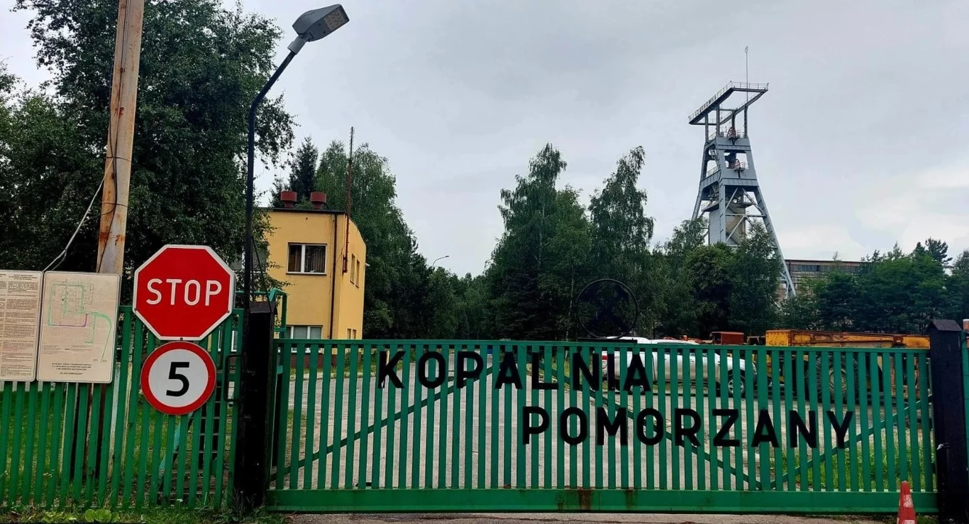 Wejście na teren kopalni Pomorzany z zieloną bramą, na której widnieje nazwa kopalni, znak STOP oraz ograniczenie prędkości do 5 km/h; w tle widoczna infrastruktura górnicza oraz drzewa.