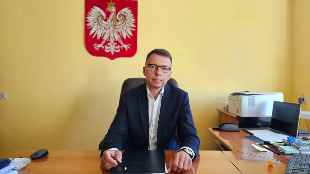 Mężczyzna w garniturze siedzi przy biurku w urzędowym gabinecie, za nim na ścianie wisi godło Polski, na biurku leżą dokumenty, komputer i drukarka.