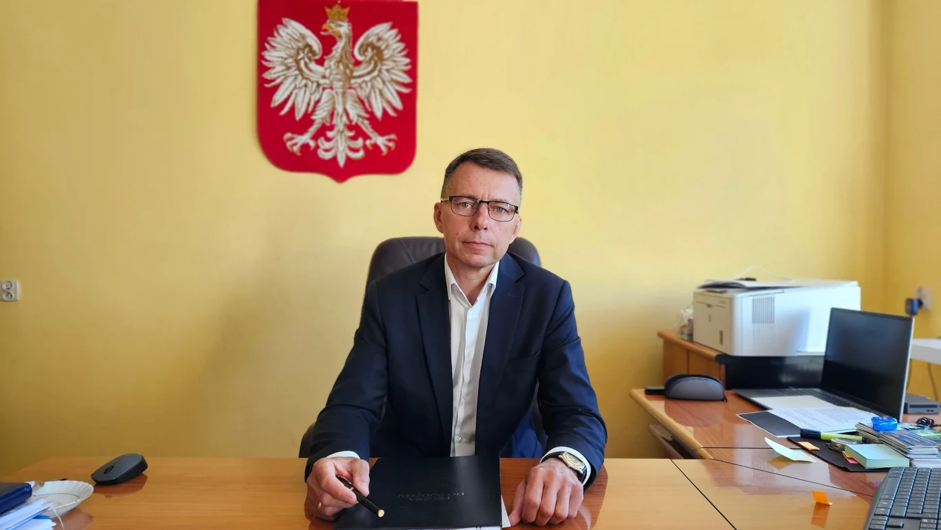 Mężczyzna w garniturze siedzi przy biurku w urzędowym gabinecie, za nim na ścianie wisi godło Polski, na biurku leżą dokumenty, komputer i drukarka.