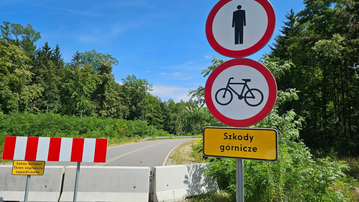 Biała betonowa bariera blokująca drogę w lesie, znaki zakazu ruchu pieszych i rowerzystów, żółta tablica informująca o szkodach górniczych, czerwono-biała tablica ostrzegawcza oraz zakaz wstępu na teren objęty zagrożeniem.