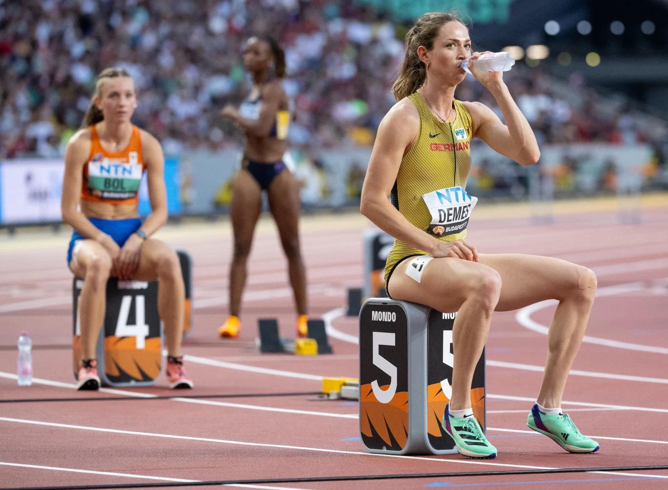 Zawodniczka w stroju sportowym siedzi na bloku startowym z numerem 5 i pije wodę, z tyłu widoczna inna sportsmenka na bloku startowym z numerem 4 oraz rozmyte sylwetki na tle tłumu kibiców na stadionie lekkoatletycznym.