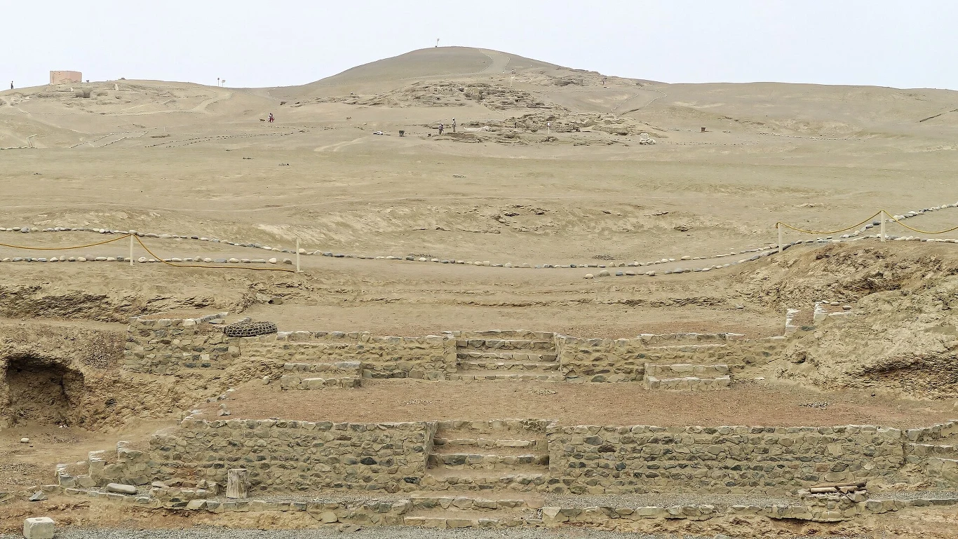 W ruinach miasta Vichama związanego z cywilizacją Norte Chico odkryto unikatową figurkę dwugłowego stworzenia. Kamienne schody i fundamenty starożytnego stanowiska archeologicznego na pustynnym terenie, otoczone barierkami i lekko pagórkowatym krajobrazem w tle.