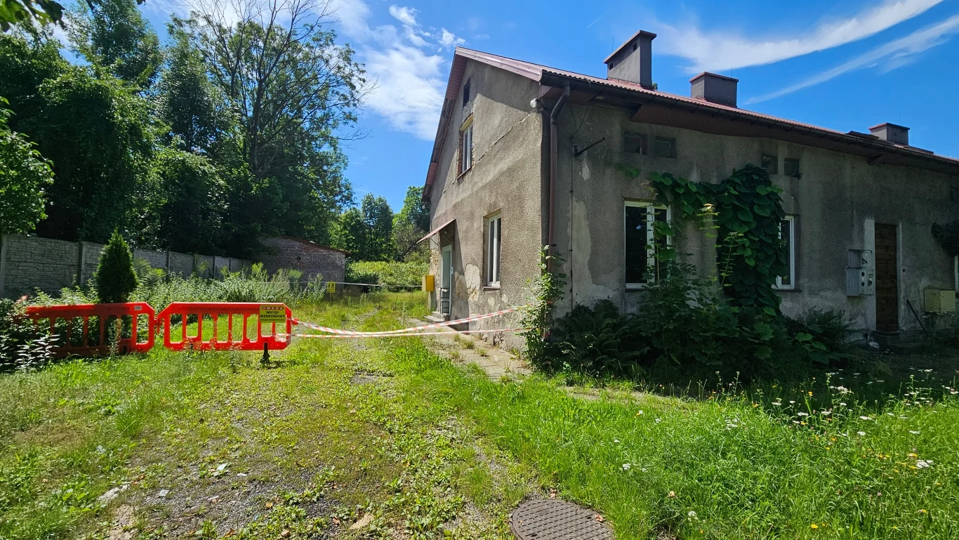 Stare, niezamieszkane domostwo otoczone wysoką zielenią, z czerwonym barierkami i taśmą ograniczającą dostęp do posesji, roślinność porastająca ściany budynku.