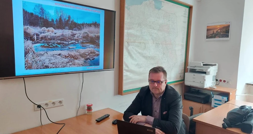 Mężczyzna siedzący przy biurku z laptopem, na ścianie za nim mapa oraz tablica z projekcją zdjęcia przyrodniczego przedstawiającego krajobraz z wodą i roślinnością; w pomieszczeniu znajduje się też drukarka i segregatory.