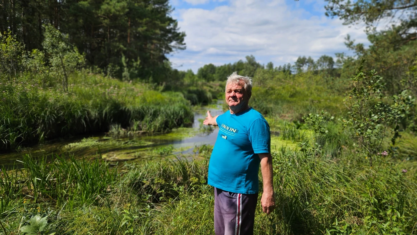 Starszy mężczyzna w niebieskiej koszulce stoi na tle zielonej łąki przy rzece, otoczonej gęstą roślinnością i drzewami, wskazując ręką w kierunku wodnego zakola.