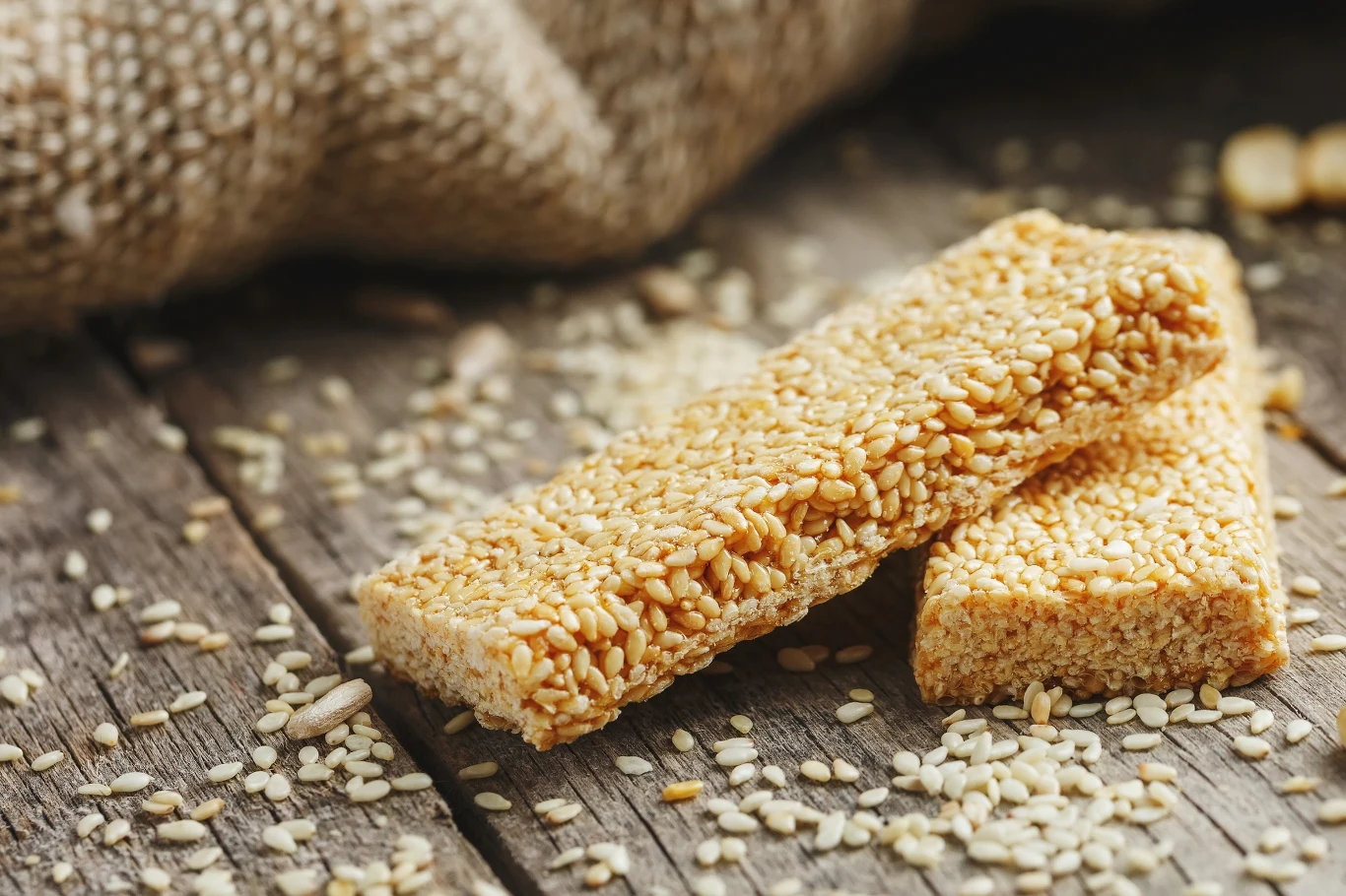 Sezamki: superfood z dawnych lat czy tylko słodka pamiątka PRL-u? Dwa batoniki sezamowe leżą na rustykalnym drewnianym stole, w otoczeniu rozsypanych ziaren sezamu, z tkaniną jutową w tle.