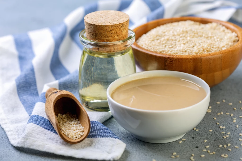 Pasta tahini w białej miseczce, słoik z olejem sezamowym, nasiona sezamu w drewnianej miseczce oraz w łyżeczce, całość ustawiona na białoniebieskiej ściereczce.