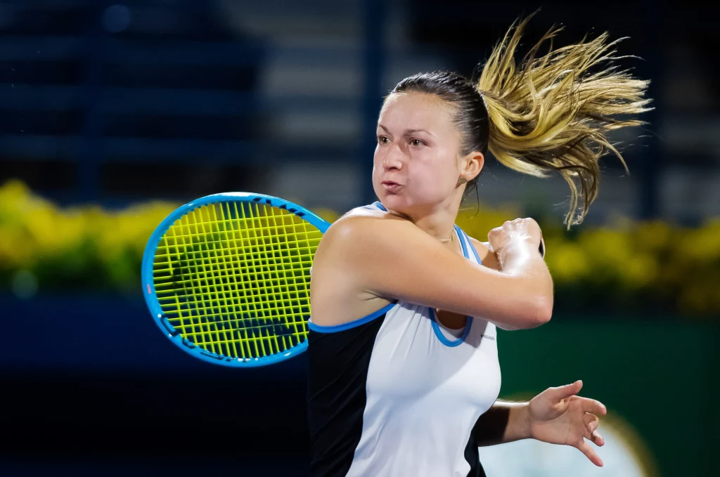 Un jugador de tenis con energía hace un intento de cohete durante un partido de tenis, en el fondo de las gradas y una fila de flores amarillas. Centrarse en la cara del jugador enfatiza la intensidad de la competencia.