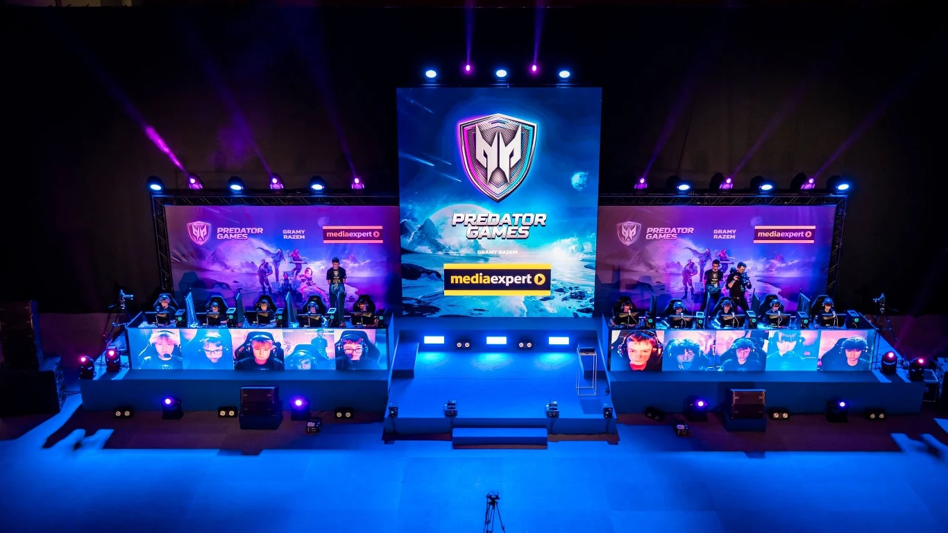 We!re Fanstasy Scena turnieju esportowego z dwoma drużynami rywalizującymi przy oddzielnych stanowiskach komputerowych, centralnie duży ekran z logo Predator Games oraz intensywne oświetlenie sceniczne. W tle widoczne banery sponsorów.