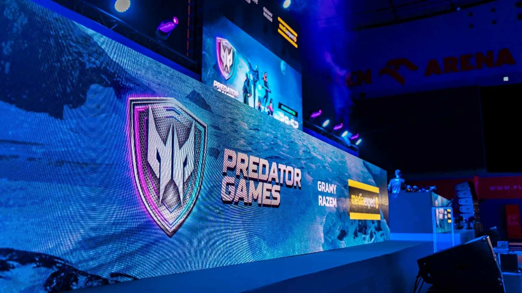 Duży podświetlony ekran z widocznym logotypem Predator Games oraz napisami i efektami świetlnymi, scena przygotowana pod wydarzenie e-sportowe z profesjonalnym oświetleniem, widoczną częścią trybun i fragmentem areny.
