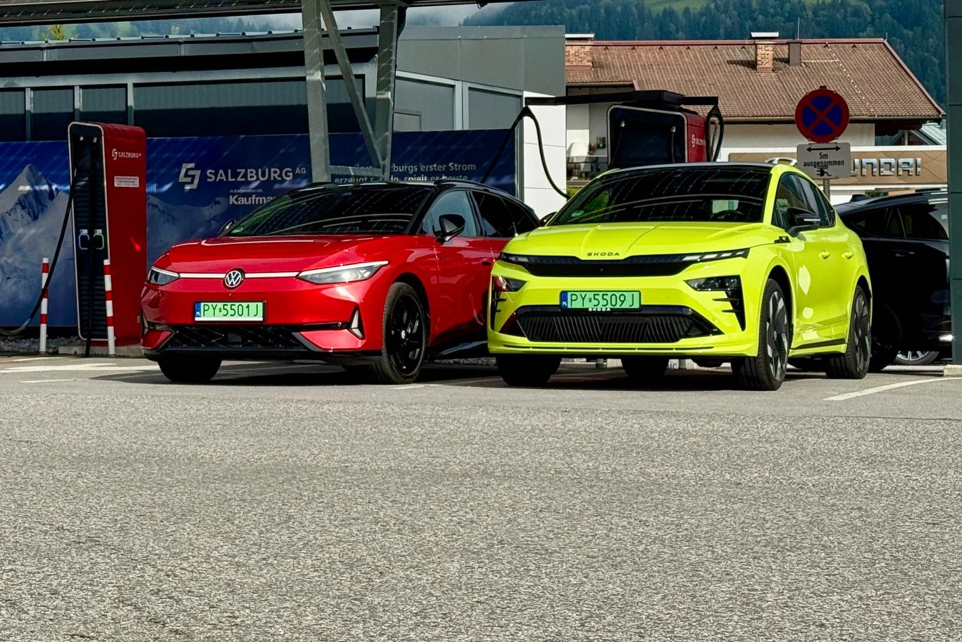 Co zrobić, gdy braknie prądu? Czy auto elektryczne można holować? Dwa nowoczesne samochody elektryczne, czerwony i żółty, zaparkowane obok siebie na stacji ładowania, oba z zielonymi tablicami rejestracyjnymi, w tle budynek i znaki drogowe.