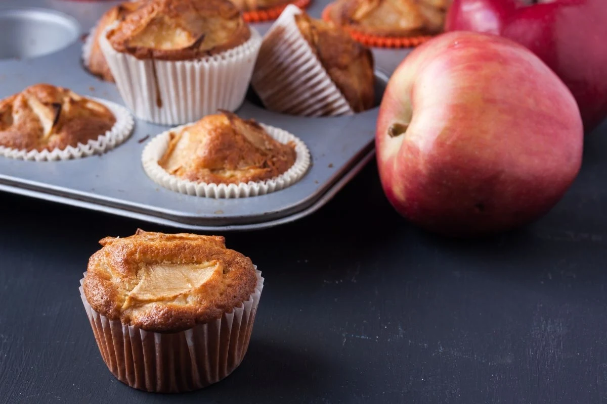 Muffinki z kawałkami jabłek w białych papilotkach ustawione w blaszce do pieczenia oraz na ciemnym stole, obok leżą czerwone jabłka.