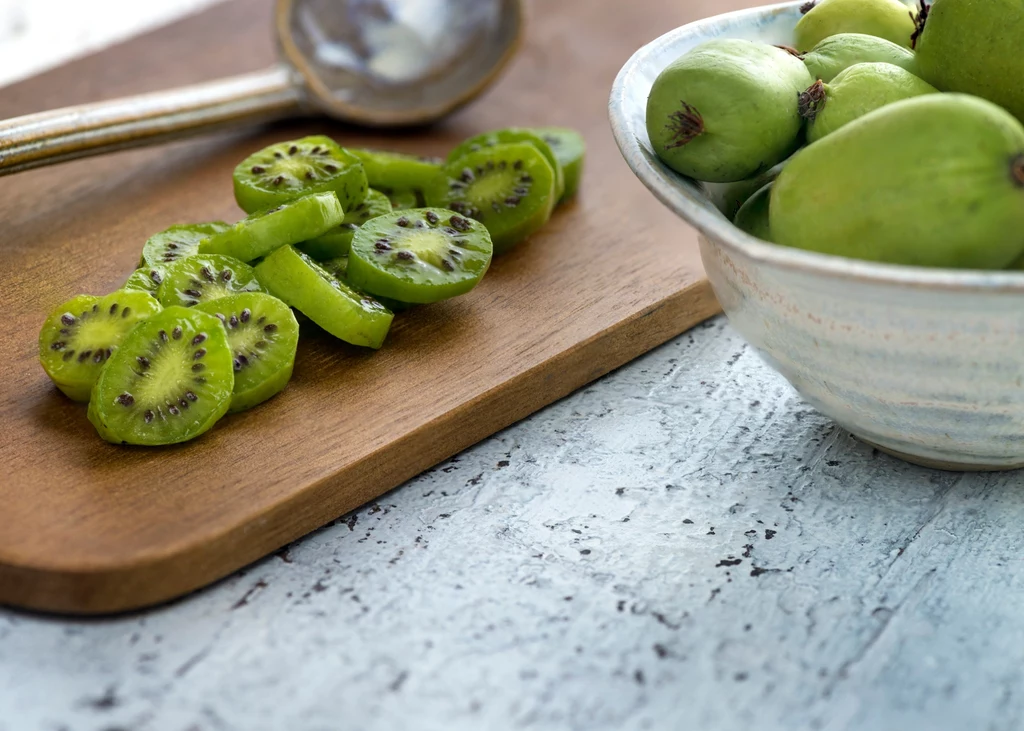 Pokrojone owoce mini kiwi na drewnianej desce obok miski z kilkoma całymi owocami, metalowa łyżka leży w tle.