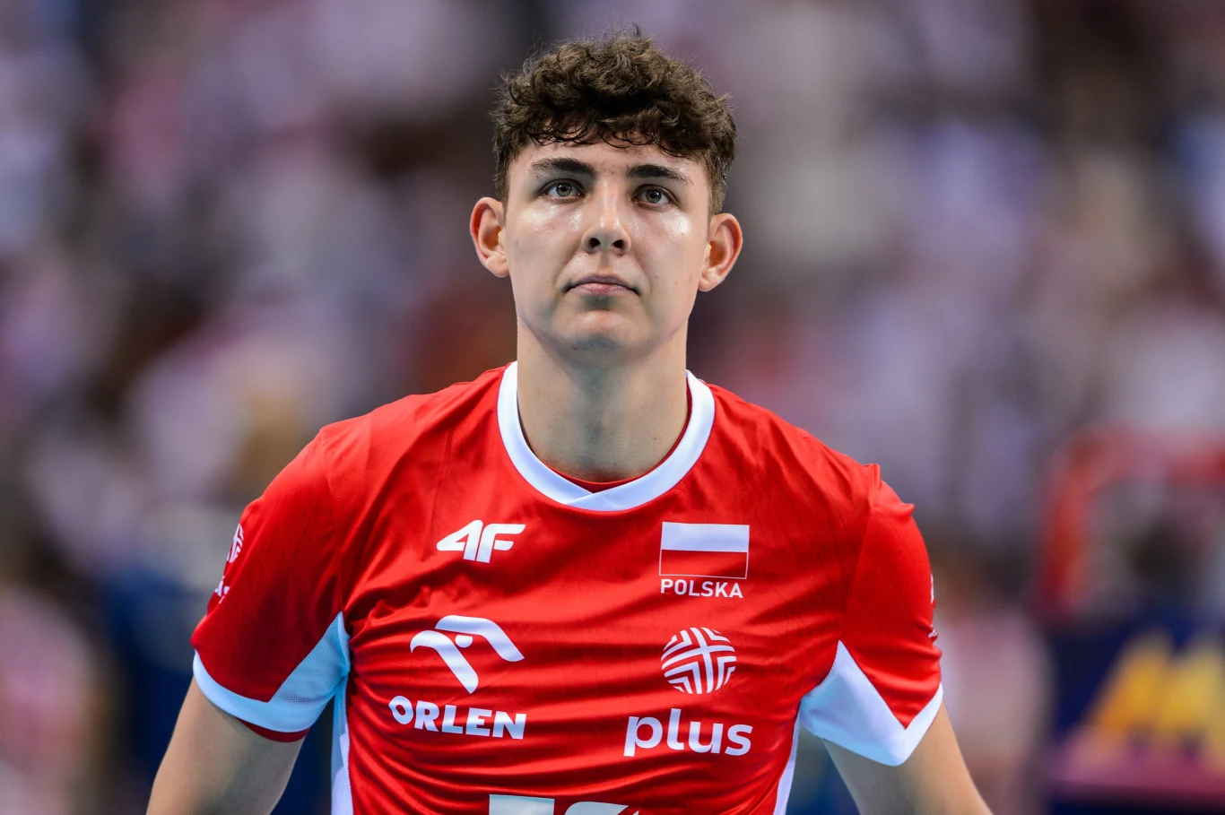 Jakub Nowak pisze piękną i nieoczywistą historię w wieku 20 lat Młody siatkarz w czerwonej koszulce reprezentacji Polski z widocznymi logotypami sponsorów i flagą kraju stoi na tle rozmytego tłumu kibiców.