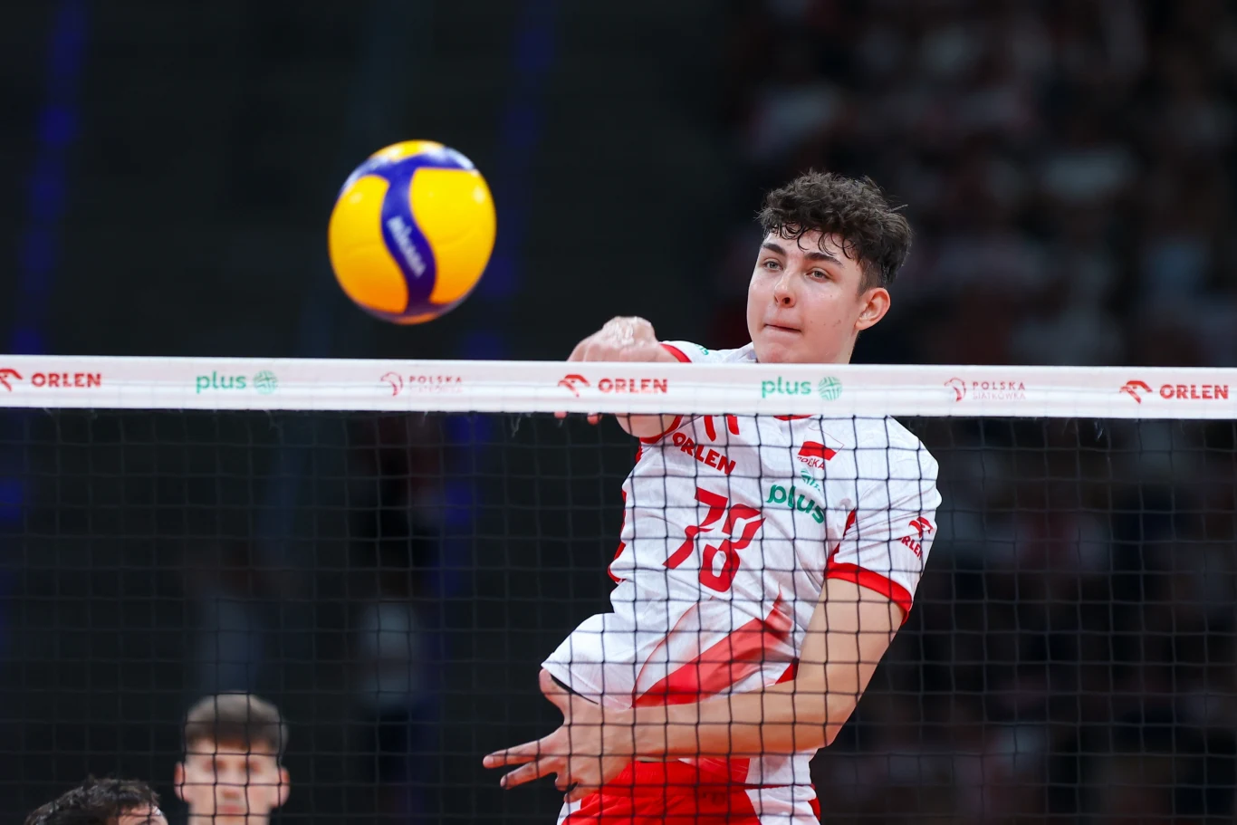 Jakub Nowak Zawodnik siatkarski w biało-czerwonym stroju wykonuje atak nad siatką, piłka unosząca się tuż nad ręką gracza, w tle widoczne rozmyte sylwetki innych osób na boisku.