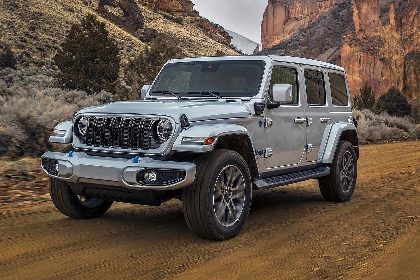 Jeep Wrangler znika z polski, To kolejna ofiara przepisów CAFE Srebrny samochód terenowy Jeep Wrangler jadący po szutrowej drodze wśród gór i skalistych wzgórz, otoczony roślinnością pustynną.