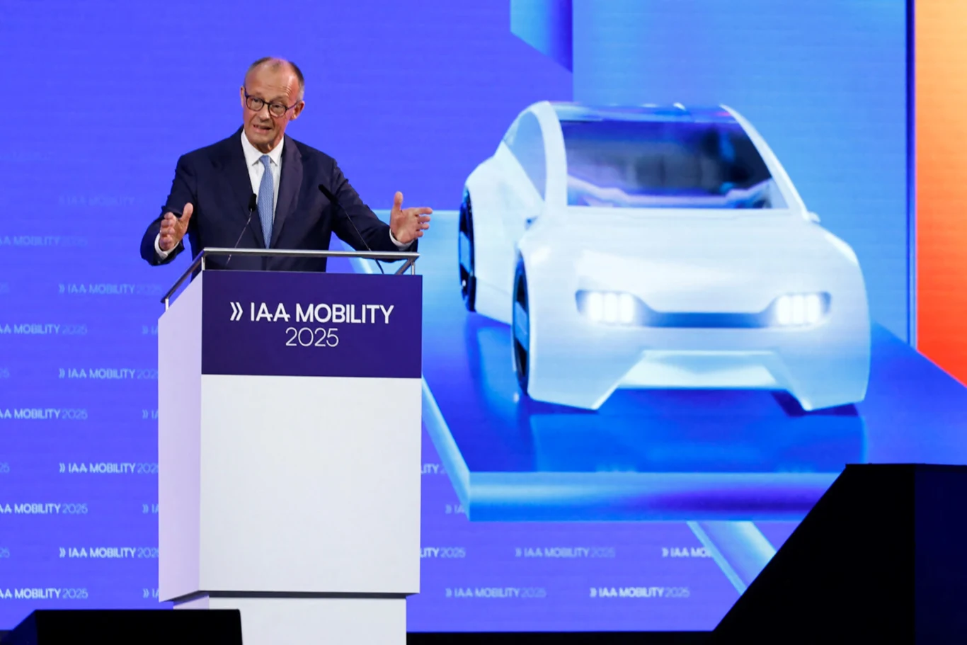 Kanclerz Niemiec z wizytą w Pekinie. Chce ratować niemiecką motoryzację Mężczyzna w garniturze przemawiający przy mównicy podczas konferencji IAA Mobility 2025, w tle znajduje się wizualizacja futurystycznego samochodu elektrycznego na dużym ekranie.