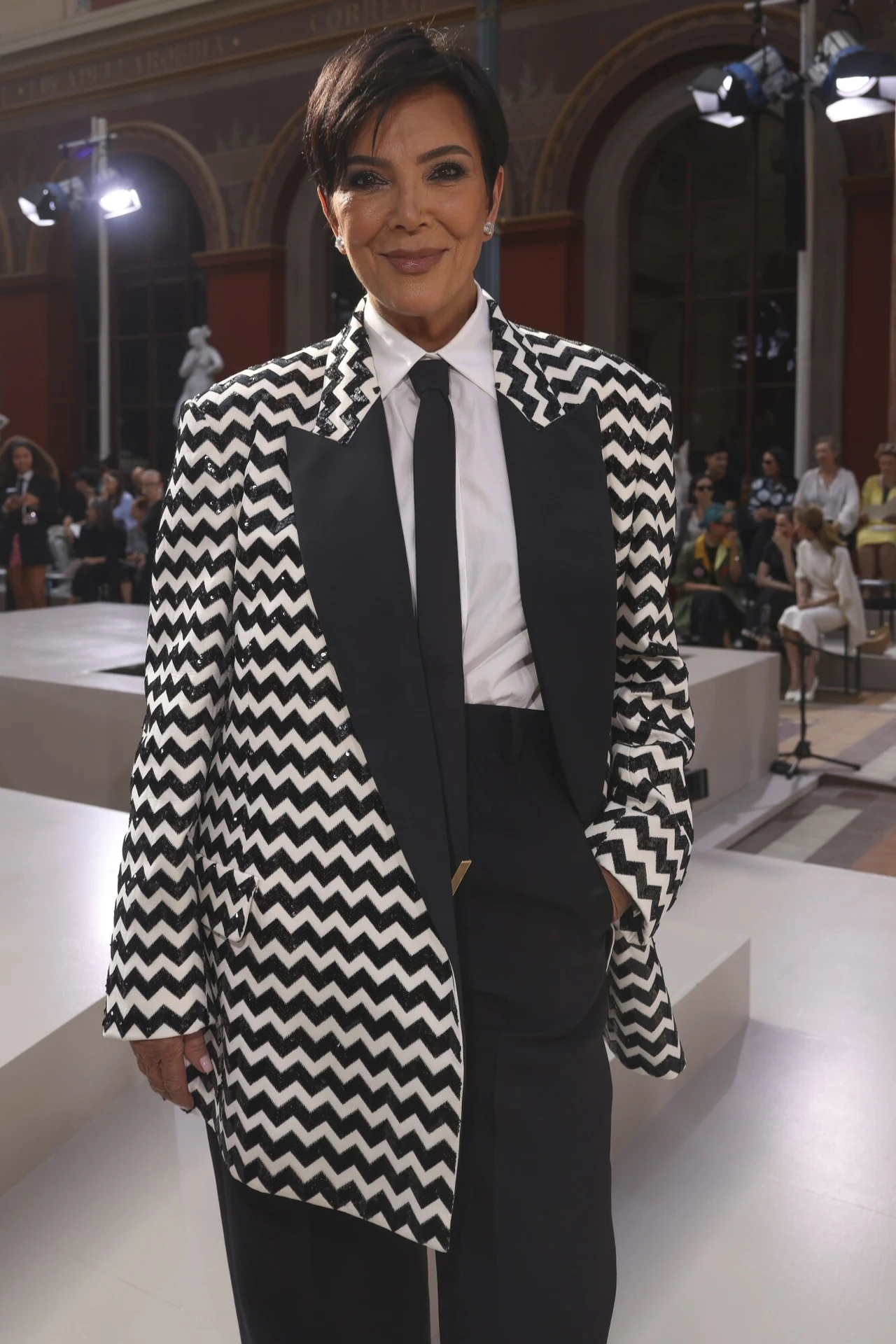 Kobieta w eleganckim stroju, szeroka marynarka w czarno-biały zygzakowaty wzór, biała koszula i czarny krawat, w tle sala z widownią i elementami architektury.