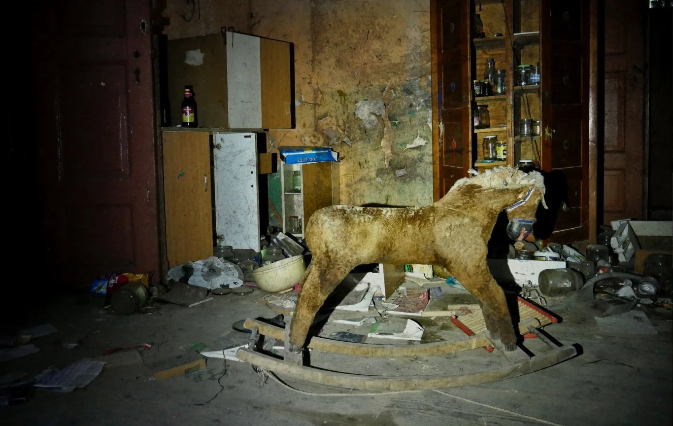 Stary, zniszczony konik na biegunach stojący na środku brudnego i zagraconego pomieszczenia, wokół rozrzucone przedmioty, puste meble i śmieci, w tle zniszczona ściana oraz otwarta szafka z porozrzucanymi przedmiotami.