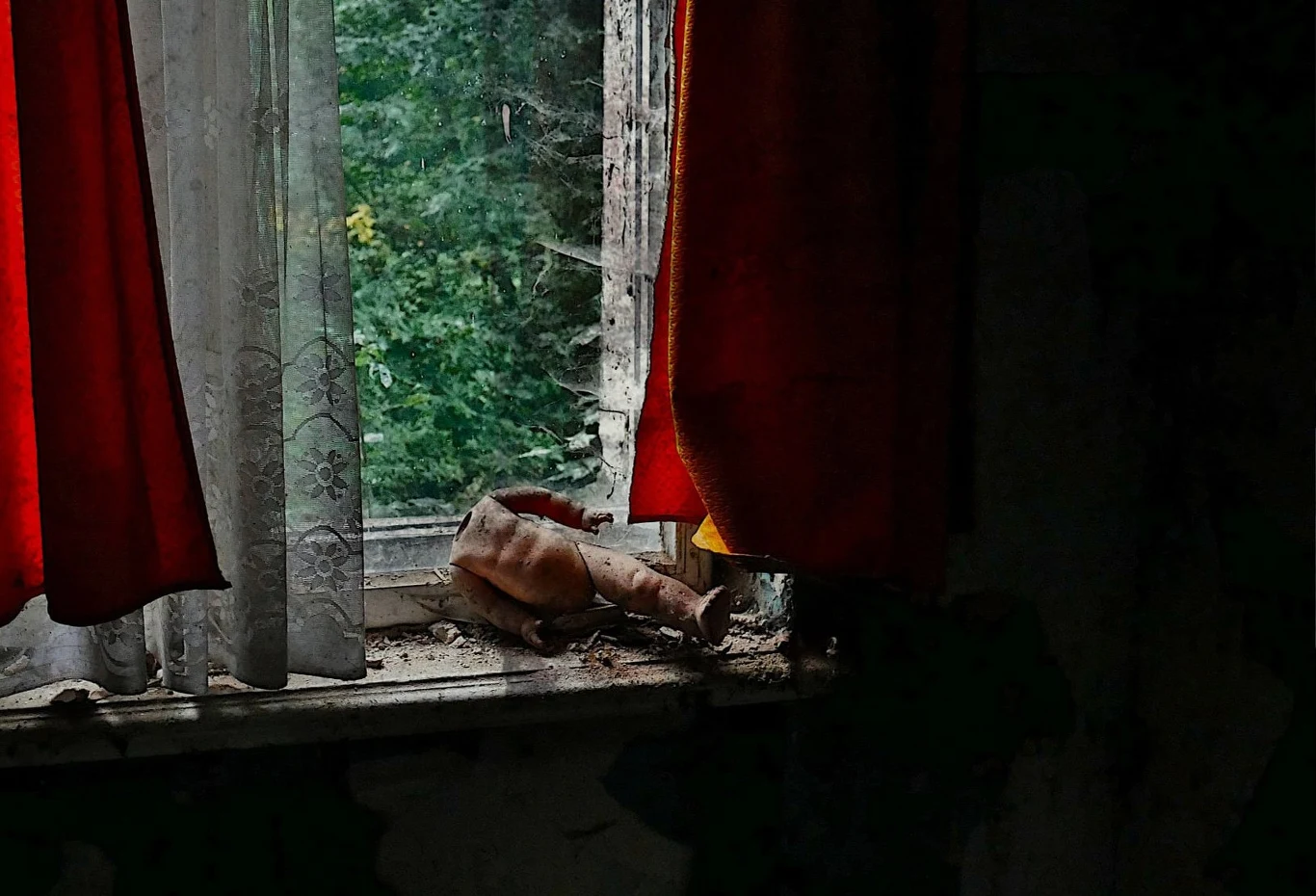Zakurzony parapet z rozdartą zabawką w opuszczonym pomieszczeniu, czerwone zasłony częściowo odsłaniają zaniedbane okno z widokiem na gęstą zieleń.