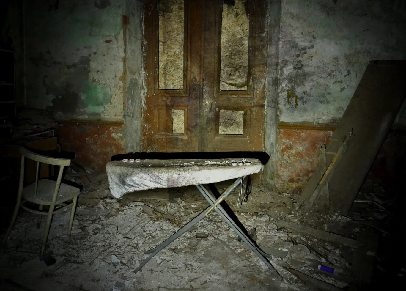 Stare, opuszczone pomieszczenie z łuszczącą się farbą na ścianach, drzwiami o widocznych śladach zniszczenia, przewrócony stolik do prasowania przykryty zabrudzonym materiałem, krzesło oraz rozrzucone gruzy na podłodze.