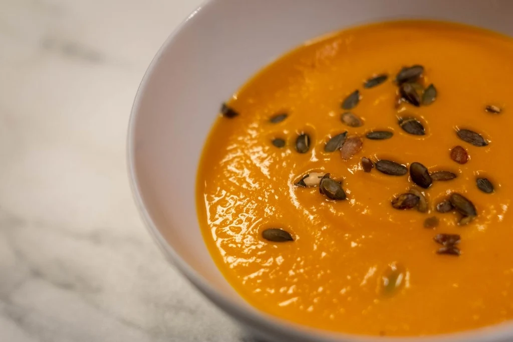 Kremowa zupa dyniowa posypana prażonymi pestkami dyni, podana w białej misce na jasnym tle.