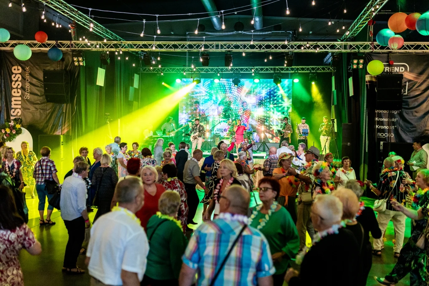 Tłum ludzi bawiący się i tańczący podczas imprezy w dużej sali, kolorowe światła sceniczne oraz dekoracje wiszące na suficie, zespół muzyczny występuje na scenie z dynamicznymi wizualizacjami w tle.