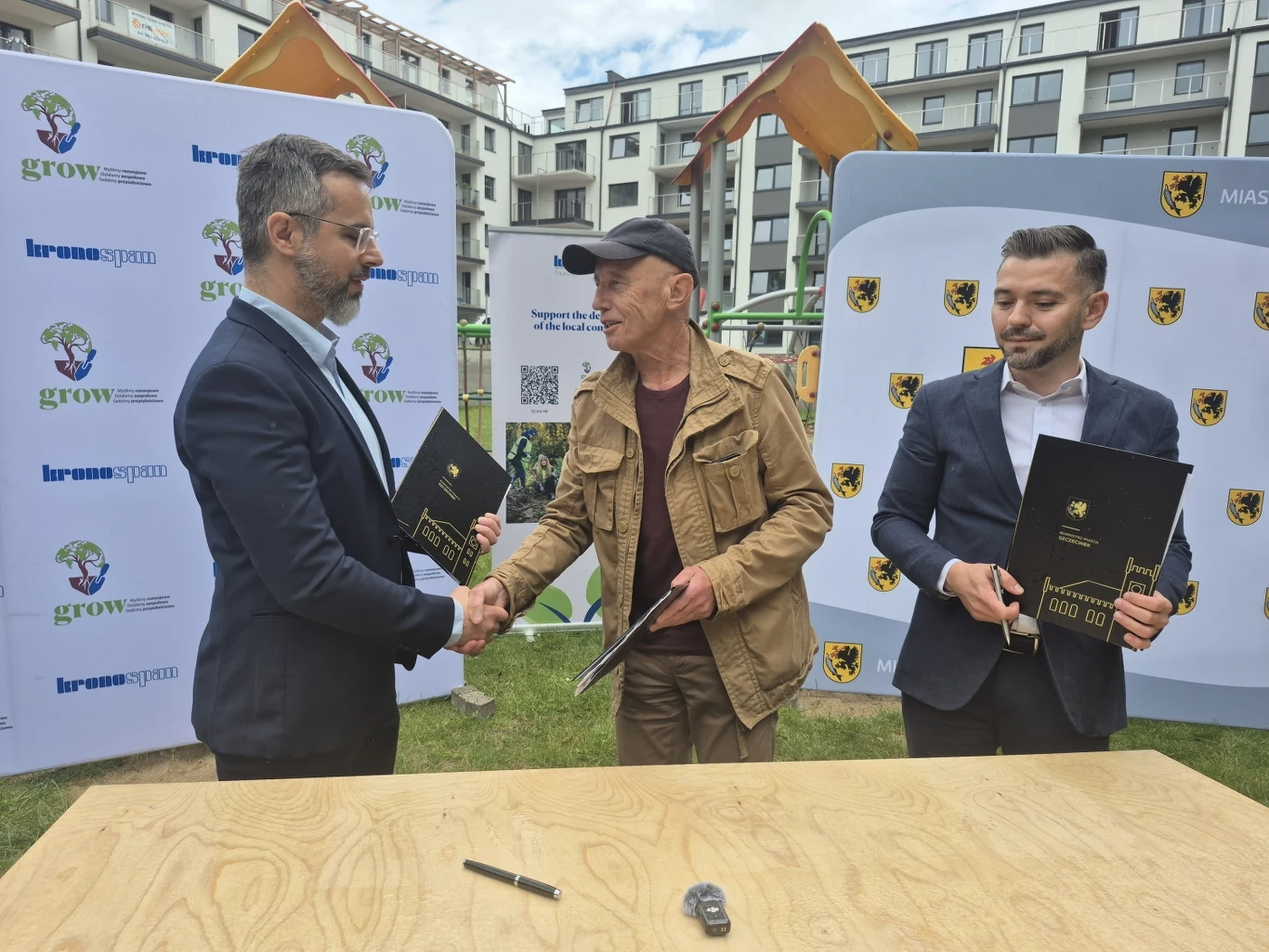 Trzech mężczyzn podczas oficjalnego spotkania na świeżym powietrzu, dwóch z nich wymienia uścisk dłoni, trzymając dokumenty, a w tle widoczne są plakaty z logotypami oraz budynki mieszkalne