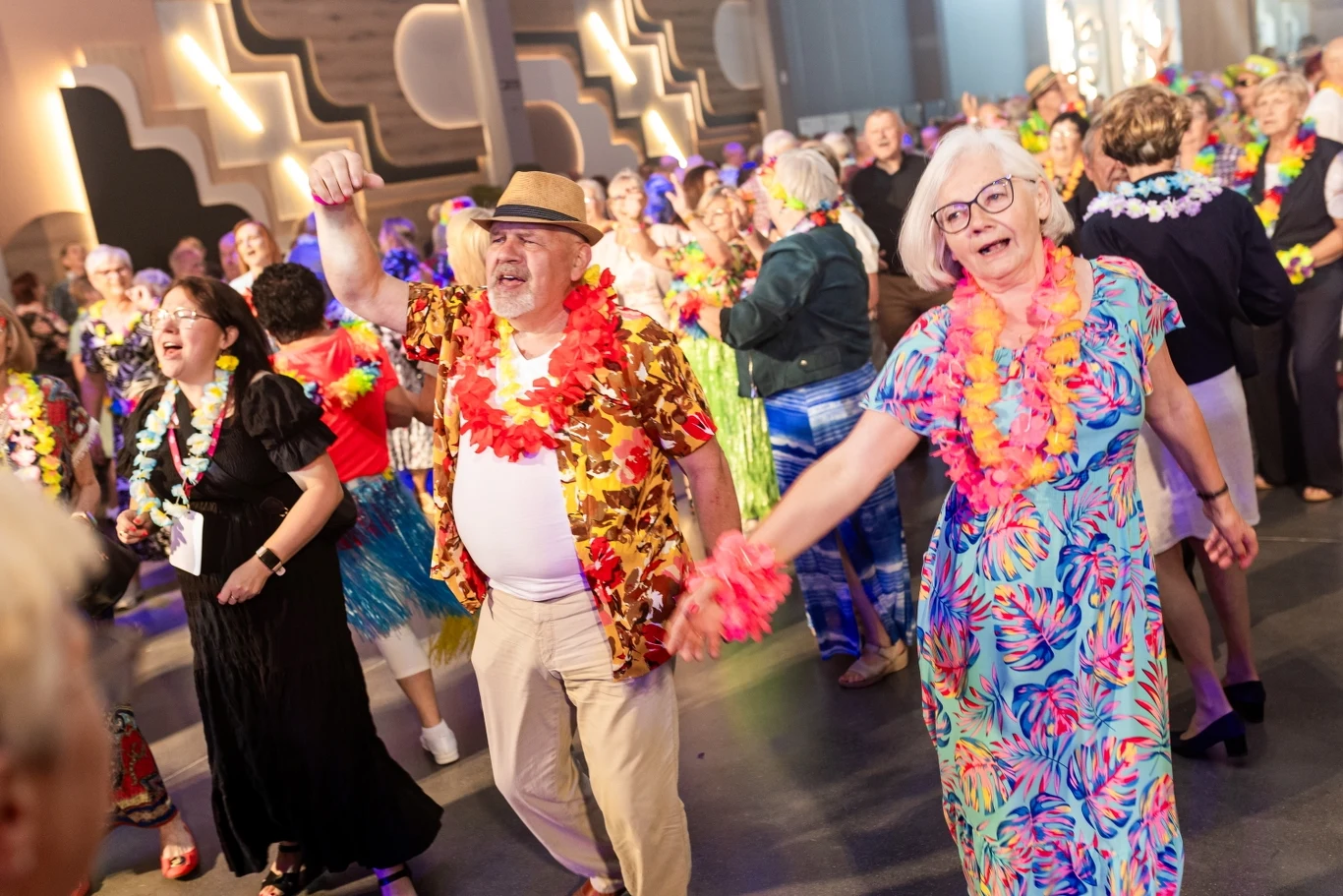 Tłum osób w kolorowych hawajskich strojach bawi się na parkiecie, starsza kobieta i mężczyzna na pierwszym planie tańczą i uśmiechają się, wokół nich panuje radosna atmosfera letniej zabawy.