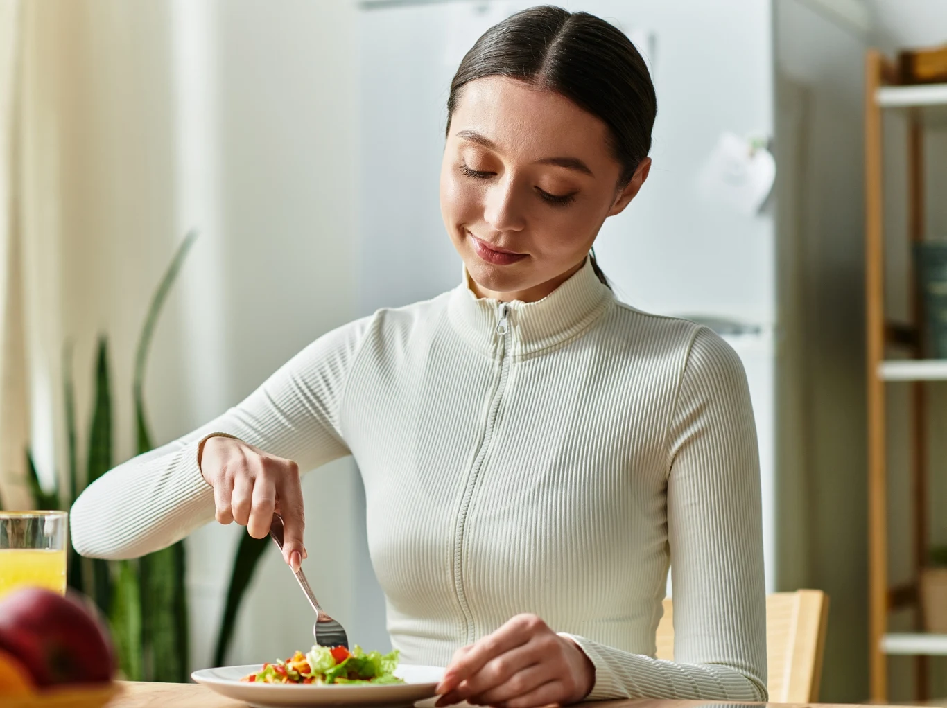 Młoda kobieta o ciemnych włosach siedzi przy stole i spożywa posiłek, na talerzu znajduje się kolorowa sałatka. Kobieta jest ubrana w białą bluzkę z długimi rękawami, w tle jasna kuchnia z lodówką oraz zieloną rośliną.