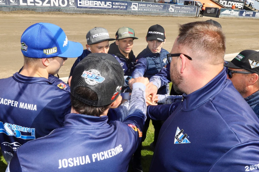 Unia Leszno Grupa zawodników w sportowych kurtkach i czapkach stojąca w kręgu z wyciągniętymi rękami, łączącymi się na środku jako symbol współpracy i jedności zespołu, tle widoczny tor żużlowy oraz reklamy na bandach.