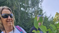 Uśmiechnięta kobieta i dziecko trzymające w rękach młode sadzonki drzew, oboje mają na sobie rękawiczki ochronne i są na tle zielonego lasu; całość wywołuje wrażenie radości i zaangażowania w akcję ekologiczną.