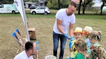 Dzieci w przebraniach oraz dorośli uczestniczą w warsztatach plenerowych, gdzie formują kulki z ziemi i wody, korzystając z misek i wiader stojących na trawie, obok stolika i koszyka z gotowymi kulami.