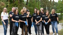Grupa ludzi w czarnych koszulkach oraz rękawiczkach ochronnych zebrana na skraju drogi przy lesie, gotowa do wspólnej pracy na świeżym powietrzu, w tle drzewa i naturalna roślinność.