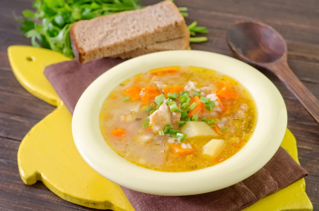 Zupa warzywna z kawałkami ziemniaków, marchewki, zieleniną i drobnymi kawałkami mięsa podana w jasnej misce, w tle kromka chleba pełnoziarnistego oraz świeża zielenina, całość ustawiona na żółtej desce i ścierce.