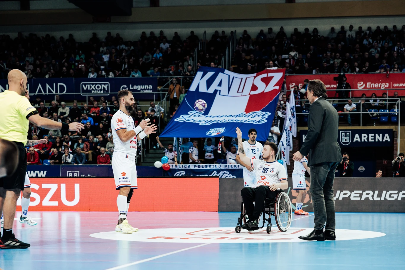 kluby ORLEN Superligi i ORLEN Superligi Kobiet wsparli kampanię pomagającą dzieciom walczącym z rakiem sportowiec na wózku inwalidzkim w białym stroju drużyny stoi na środku hali sportowej, wokół niego inni sportowcy oraz sędzia, na tle trybun powiewa duża flaga z napisem 'Kalisz', atmosfera wydarzenia sportowego z udziałem licznej publiczności
