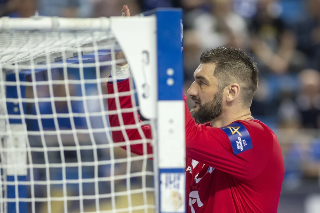 Bramkarz ubrany w czerwony strój sportowy stoi blisko słupka bramki podczas meczu piłki ręcznej, skupiając się na sytuacji przed sobą, w tle widoczne rozmazane trybuny.