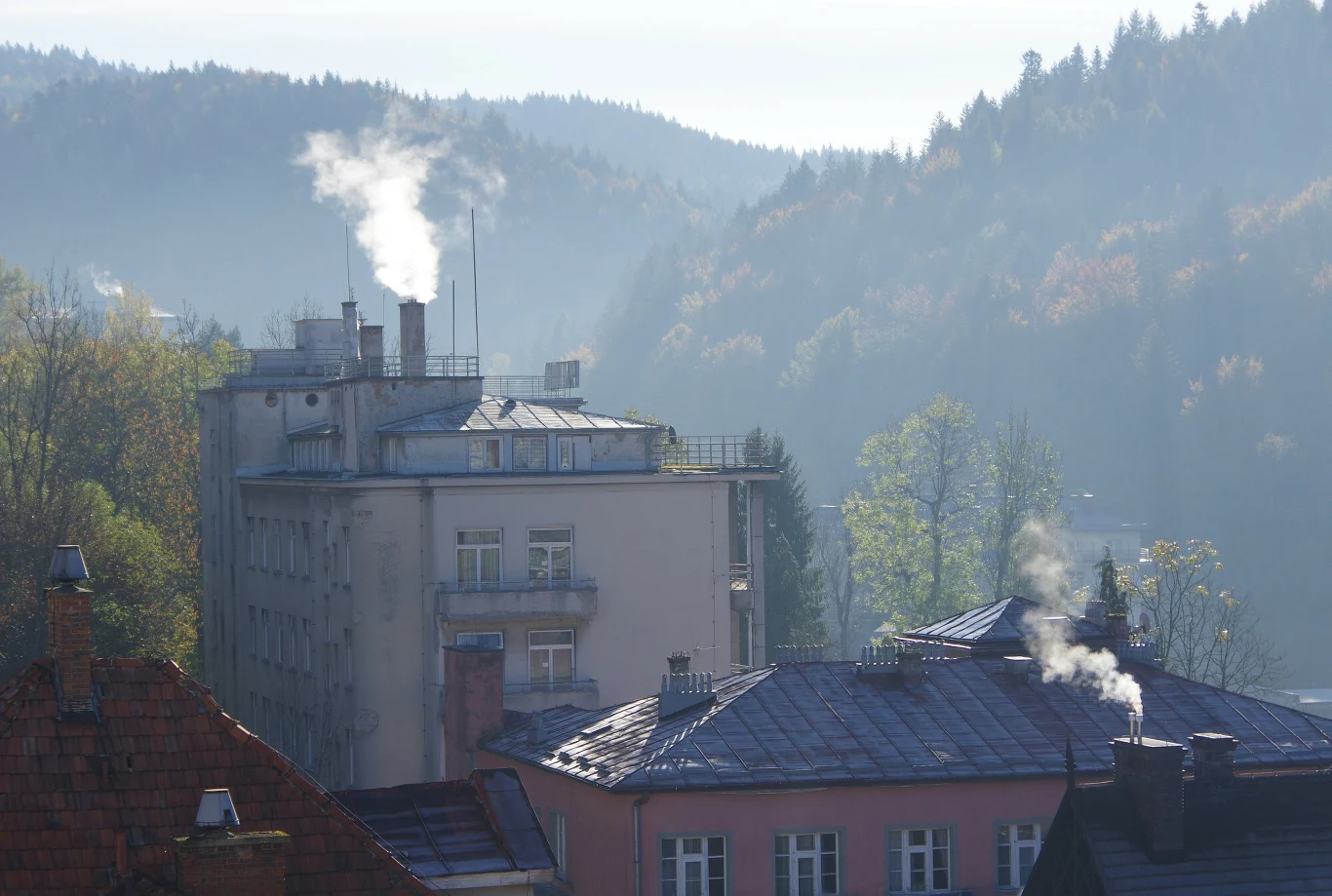 Program "Czyste Powietrze" w kryzysie. Spadek wniosków o 80 proc. (zdj. ilustracyjne) Dachy kamienic z kominami, z których unosi się dym, w tle budynek mieszkalny oraz zalesione wzgórza, delikatna mgła tworząca nastrojowy klimat.