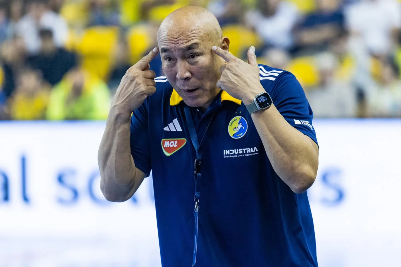Trener drużyny sportowej w niebieskiej koszulce przemawia gestykulując dłońmi przy głowie, na tle rozmytej publiczności i żółtych siedzisk.