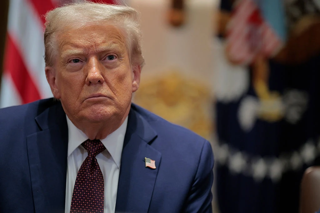 Rosyjskie drony nad Polską. Doland Trump zabrał głos Mężczyzna w garniturze i białej koszuli z poważnym wyrazem twarzy, na piersi przypięta flaga USA, w tle widoczna flaga amerykańska i elementy oficjalnego wystroju.